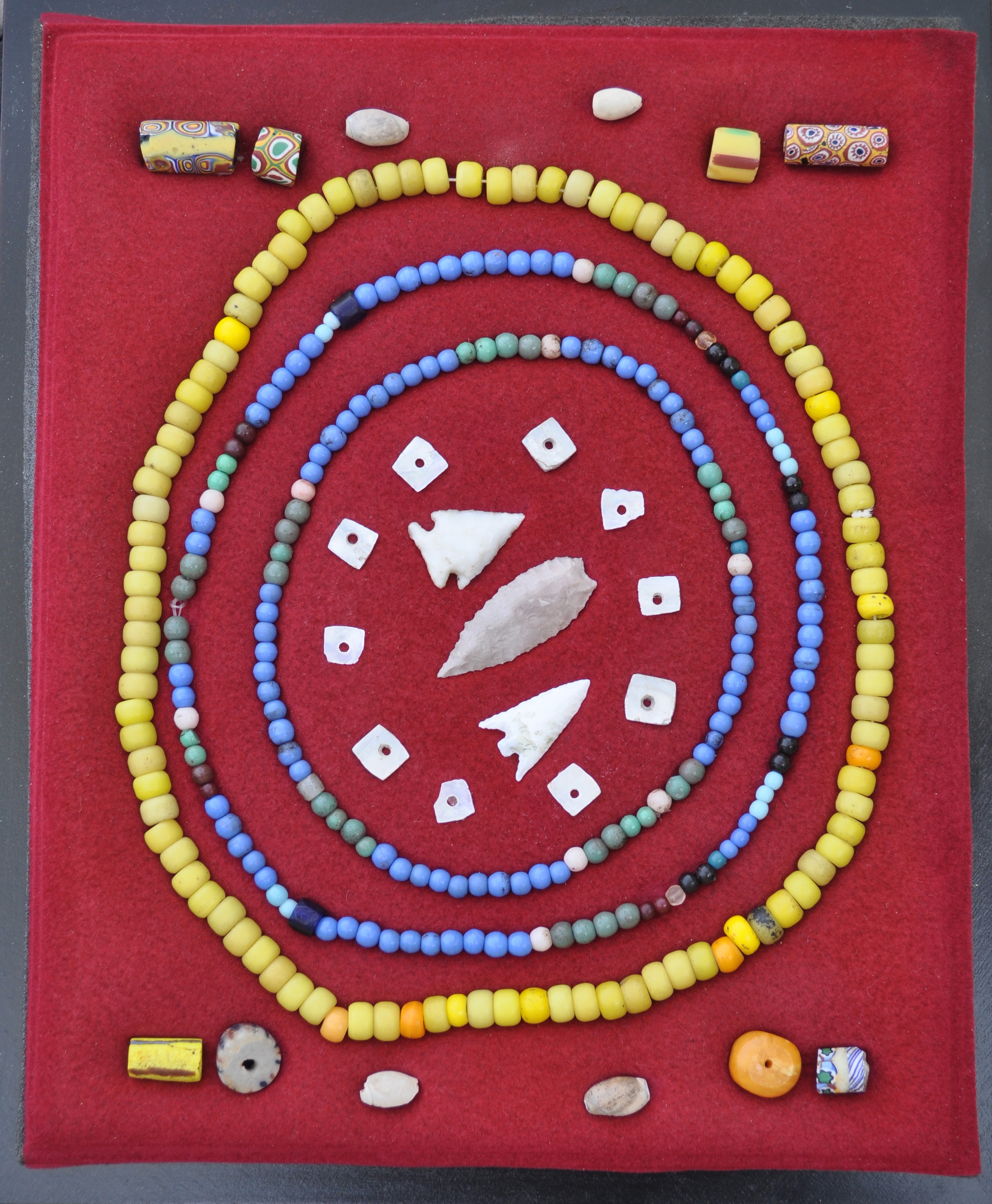 Frame with African trade beads, shell beads, padre beads, Prosser molded beads, mother-of- pearl beads, and arrow points on a red background.